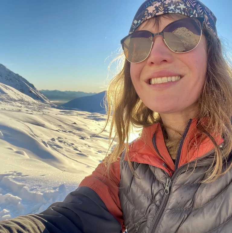 Amanda Sassi in sunglasses, a patterned headband and an orange jacket smiles on a sunlit snowy slope.