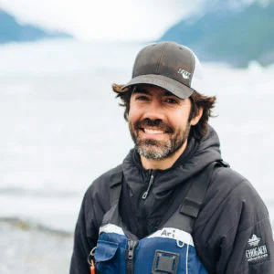Ari Stiassny smiles in a cap and a blue life vest with his name on it, a glacier blurred behind him.