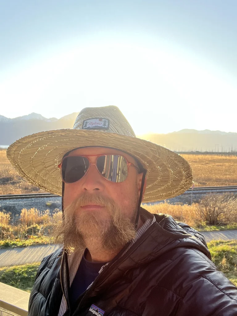 Ben Kohler, bearded, in a straw hat and sunglasses takes a selfie with low sun, rail tracks and mountains behind.