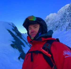 Brian Burnett in a red jacket, helmet and goggles, wearing a pack below snowy peaks and a blue sky.