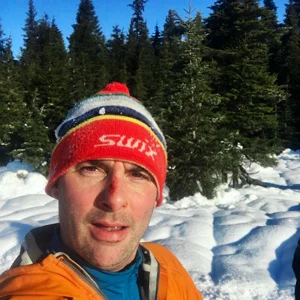 Brian Kari in a red Swix hat and orange jacket takes a selfie among snowy spruce trees under a blue sky.