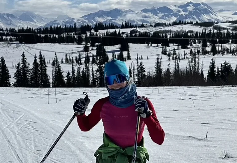 Briana Sullivan in a teal hat, goggles and red top skis on a groomed trail, snowy mountains behind.