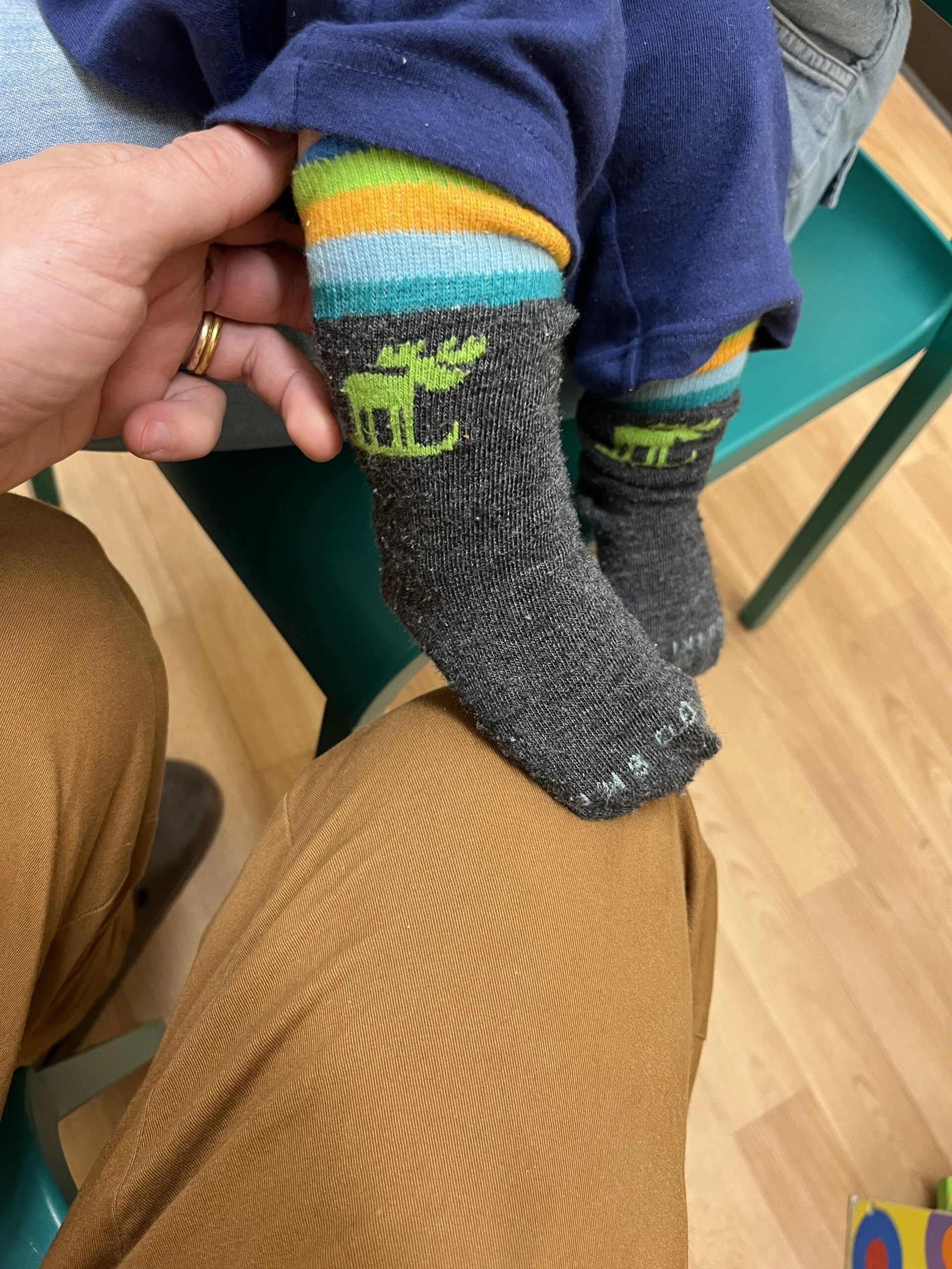 Small child in gray socks with green moose and rainbow-striped cuffs, sitting on an adult's lap.