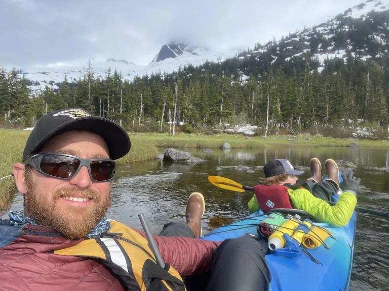 Chris Stinson takes a selfie in a blue tandem kayak with a paddler up front, spruce and snowy peaks behind.