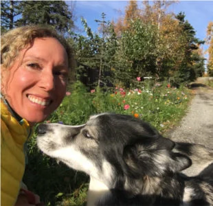 Christina Gerber in a yellow jacket smiles beside a grey and white husky by wildflowers and autumn trees.