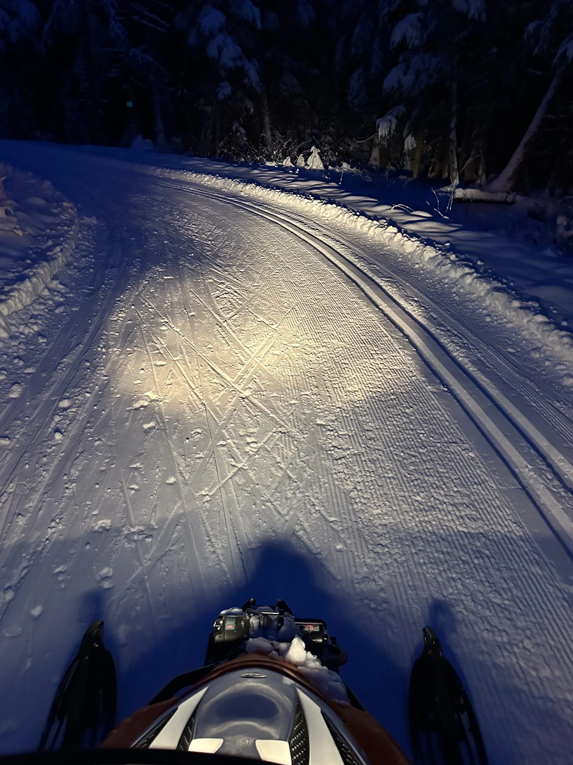 From a snowmachine at night, the headlight shows a curving trail with classic tracks and ski marks in the snow.