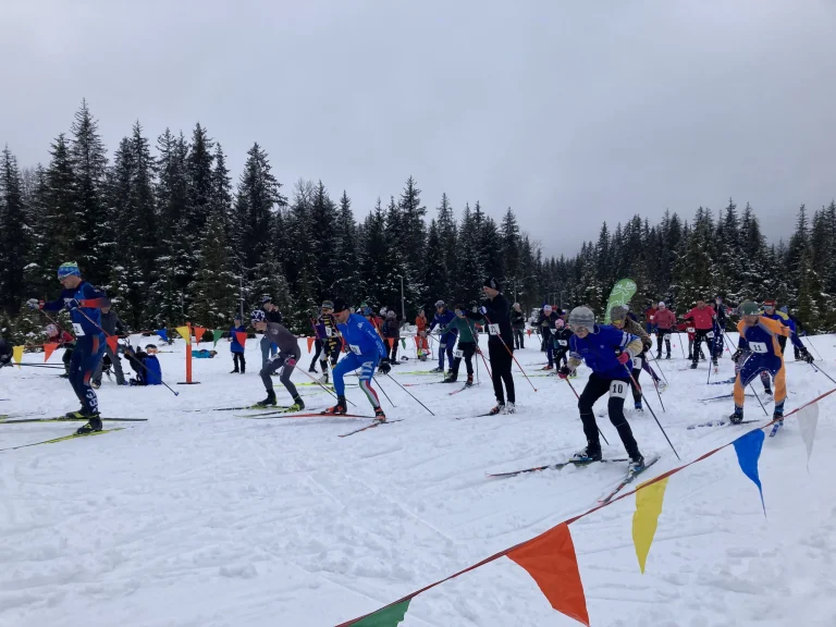 Racers in bibs push off from a mass start on snow behind a string of colorful pennant flags, with spruce forest beyond.