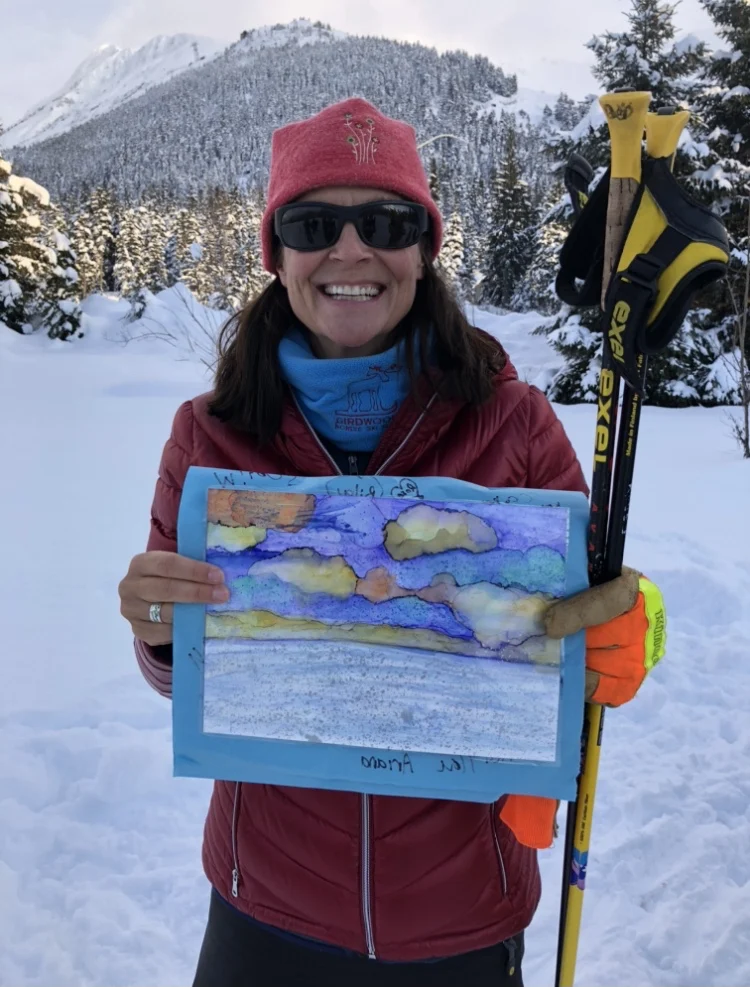 Deb Essex in a red jacket and hat smiles holding a painted sky scene and ski poles, snowy peaks behind.