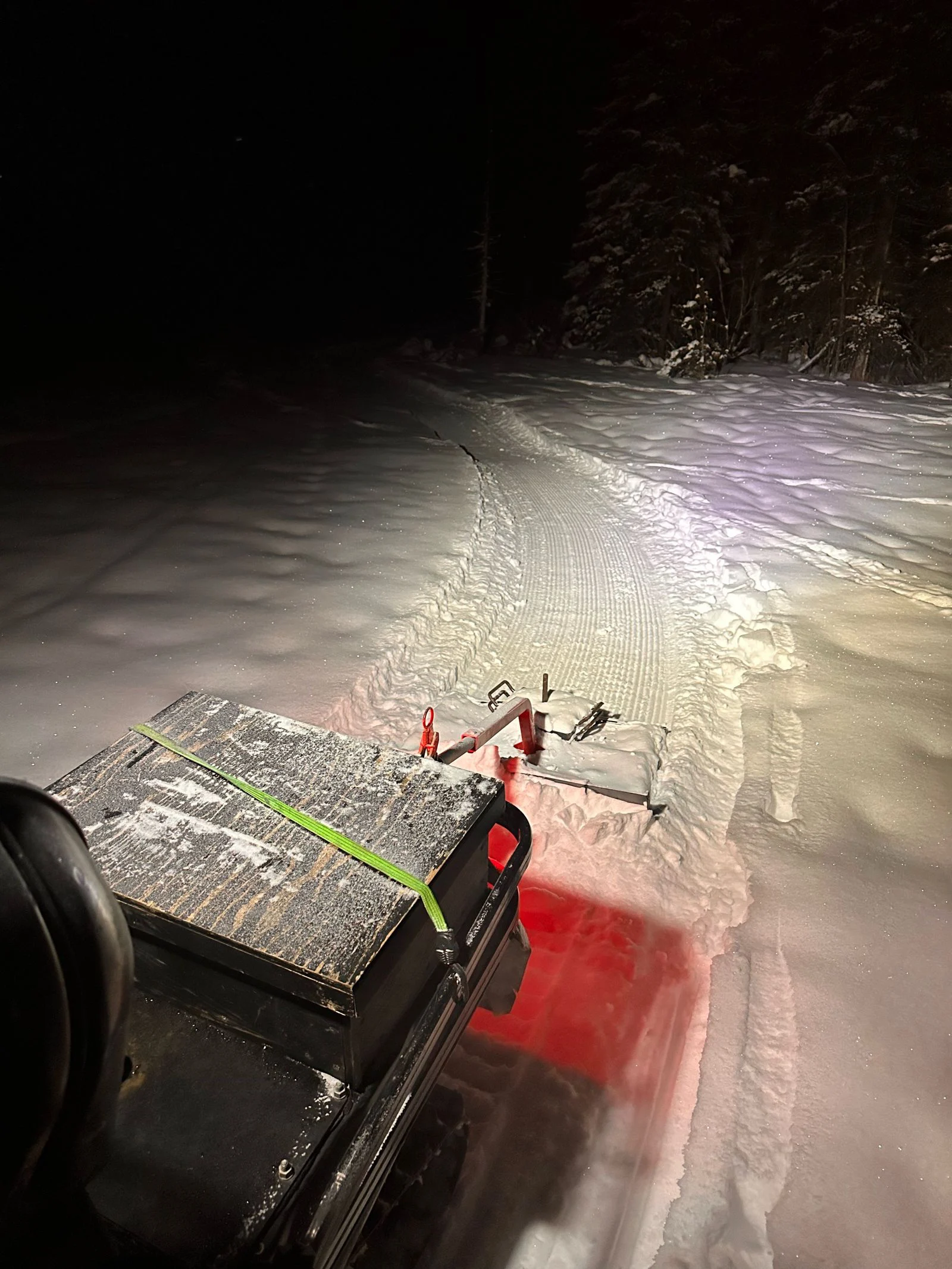 Night view back over a snowmachine's strapped cargo box to a small drag leaving narrow corduroy through deep snow.
