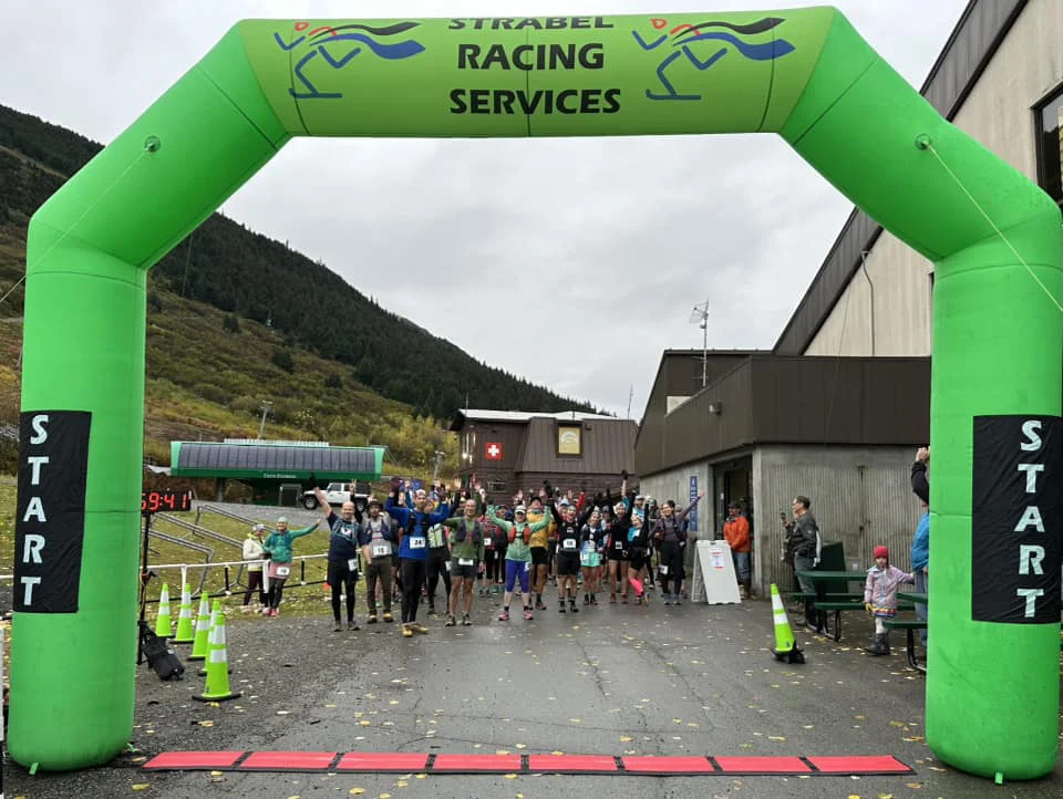 Runners raise their arms under a green inflatable Strabel Racing Services start arch on a cloudy autumn day.