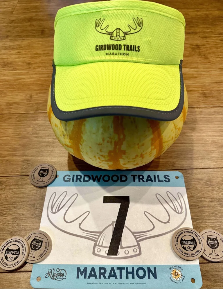 Neon yellow Girdwood Trails Marathon visor on a striped squash, above race bib number 7 and wooden beer tokens.