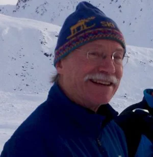 Jim Braham, with glasses and a white mustache, smiles in a blue knit ski hat in front of a snowy slope.