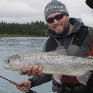 Justin Thomas in a beanie and sunglasses smiles holding a large rainbow trout beside a river.
