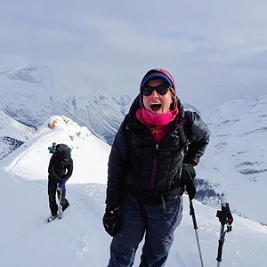 Karen Loso in a pink beanie and black puffy jacket laughs on a snowy ridge as another person climbs behind.