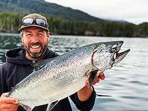 Nels Barber in a cap and dark hoodie grins holding a large silver salmon on the water, forested hills behind.