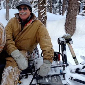 Peter Zug in a tan work jacket, knit hat and sunglasses sits on a snow-covered snowmachine among spruce trees.
