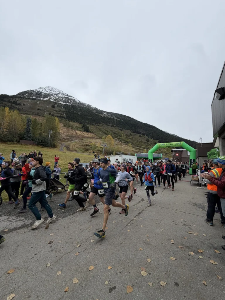 Runners leave the start under a green arch at the Girdwood Marathon, a snow-dusted peak behind them.