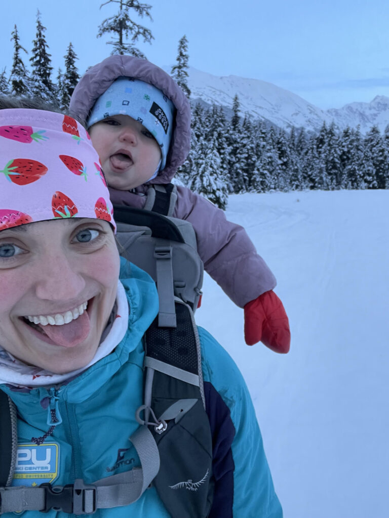 Lauren Fritz smiles in a selfie on a snowy trail, with a baby in a backpack carrier behind her.