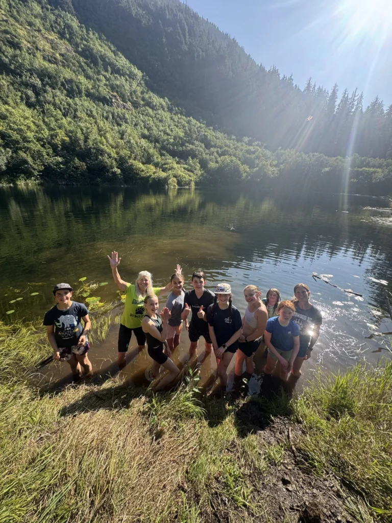 Girdwood middle school cross country runners wave from the grassy shore of a sunlit mountain lake.